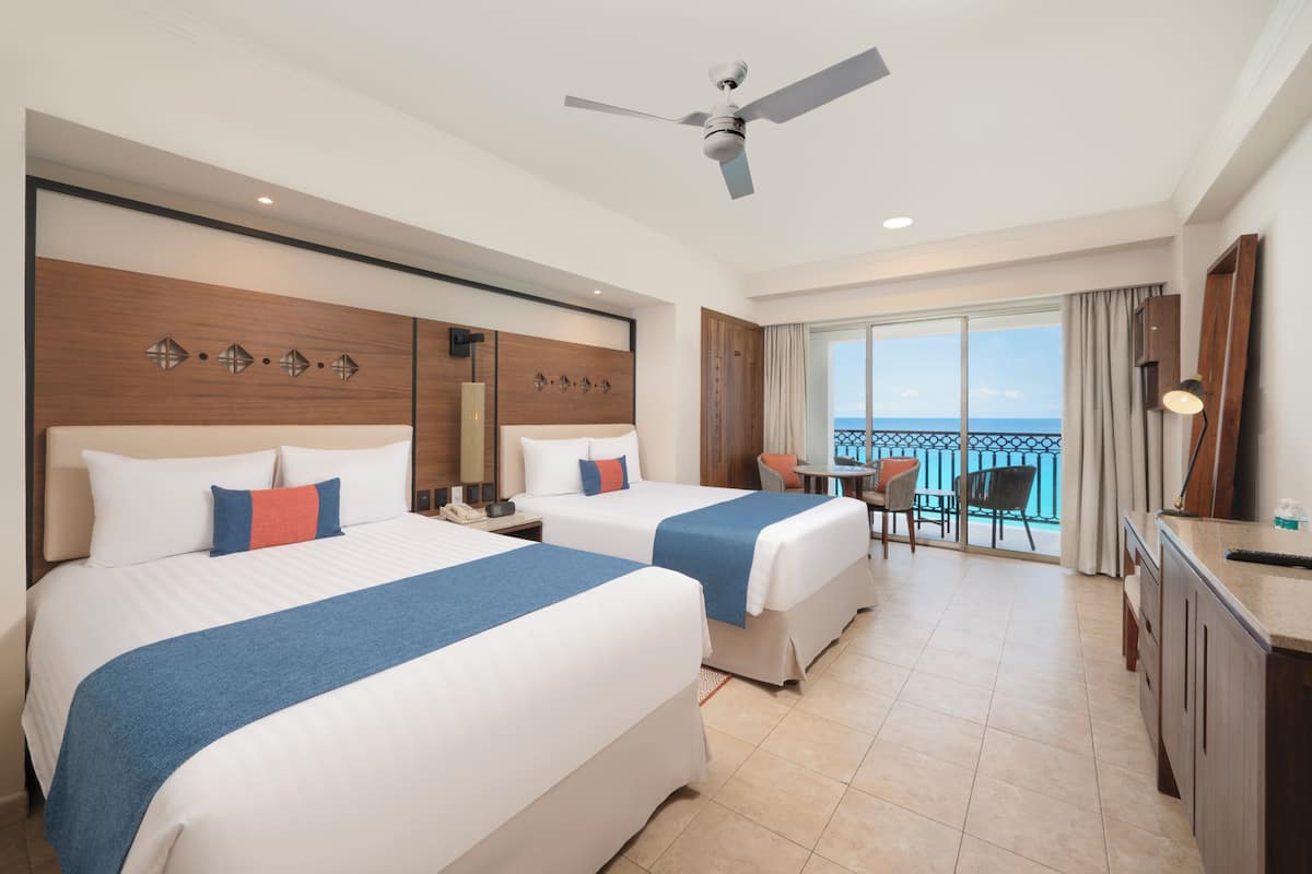A hotel room with two beds, a balcony with a view, a desk, and a ceiling fan.