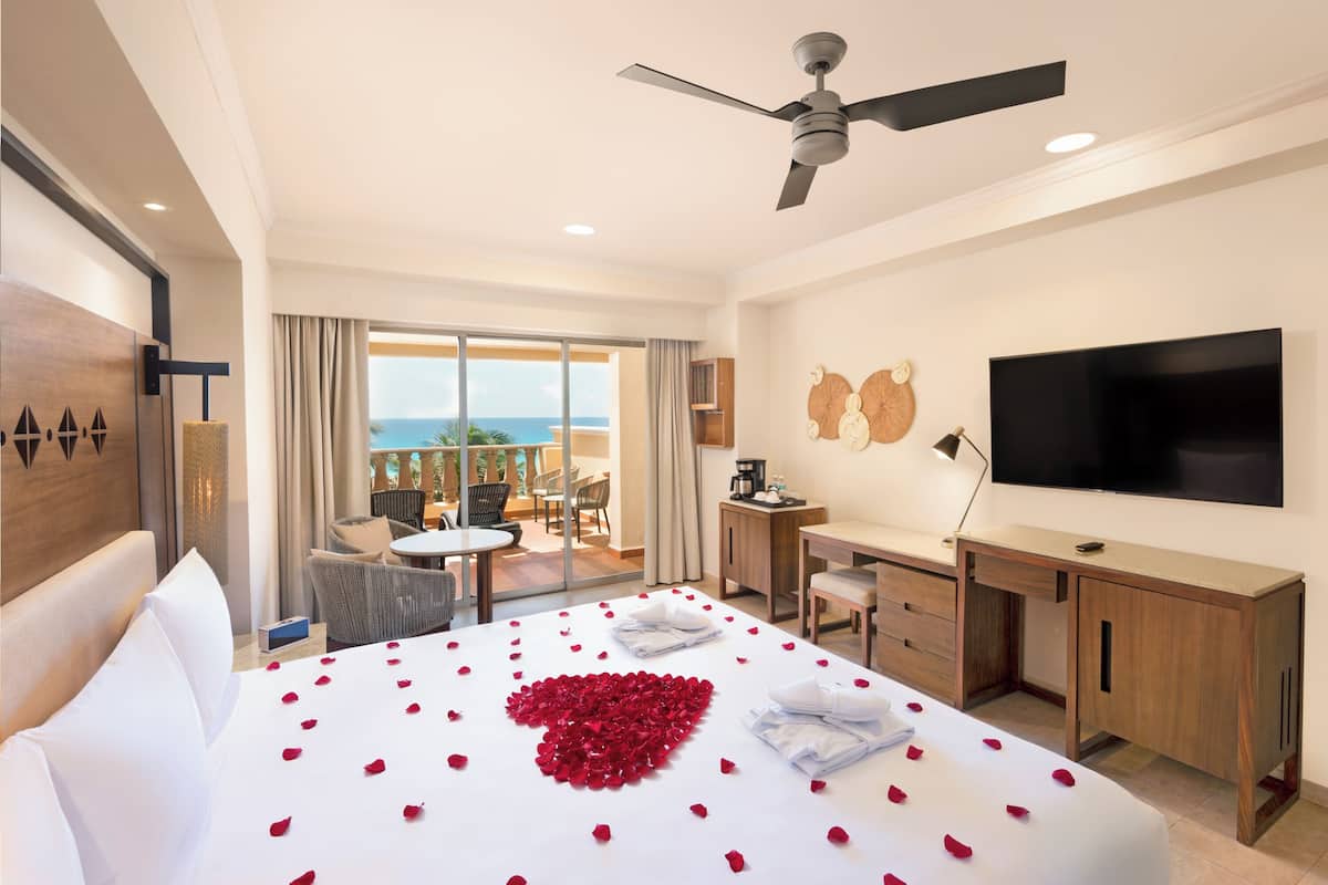 A hotel room with a bed, a desk, a TV, and a balcony with a view of the ocean.