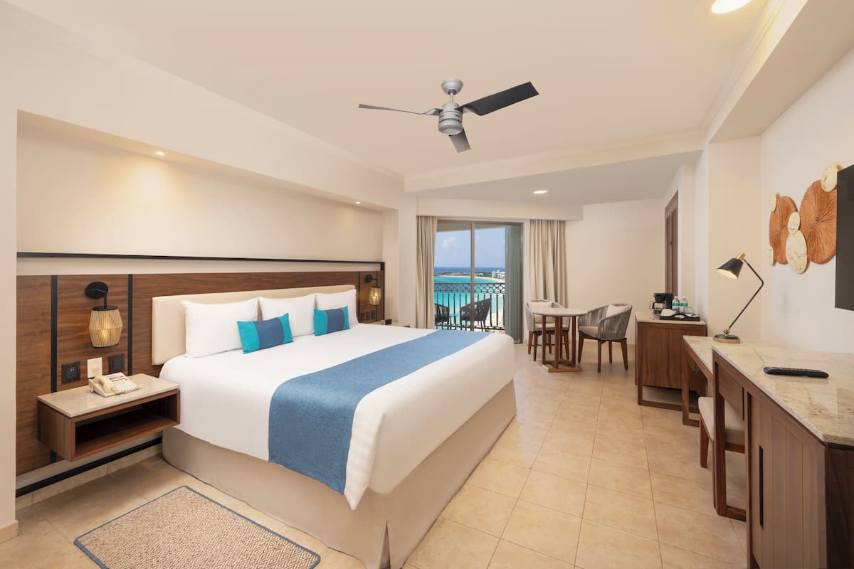 A hotel room with a large bed, a desk, a chair, a ceiling fan, and a view of the ocean.