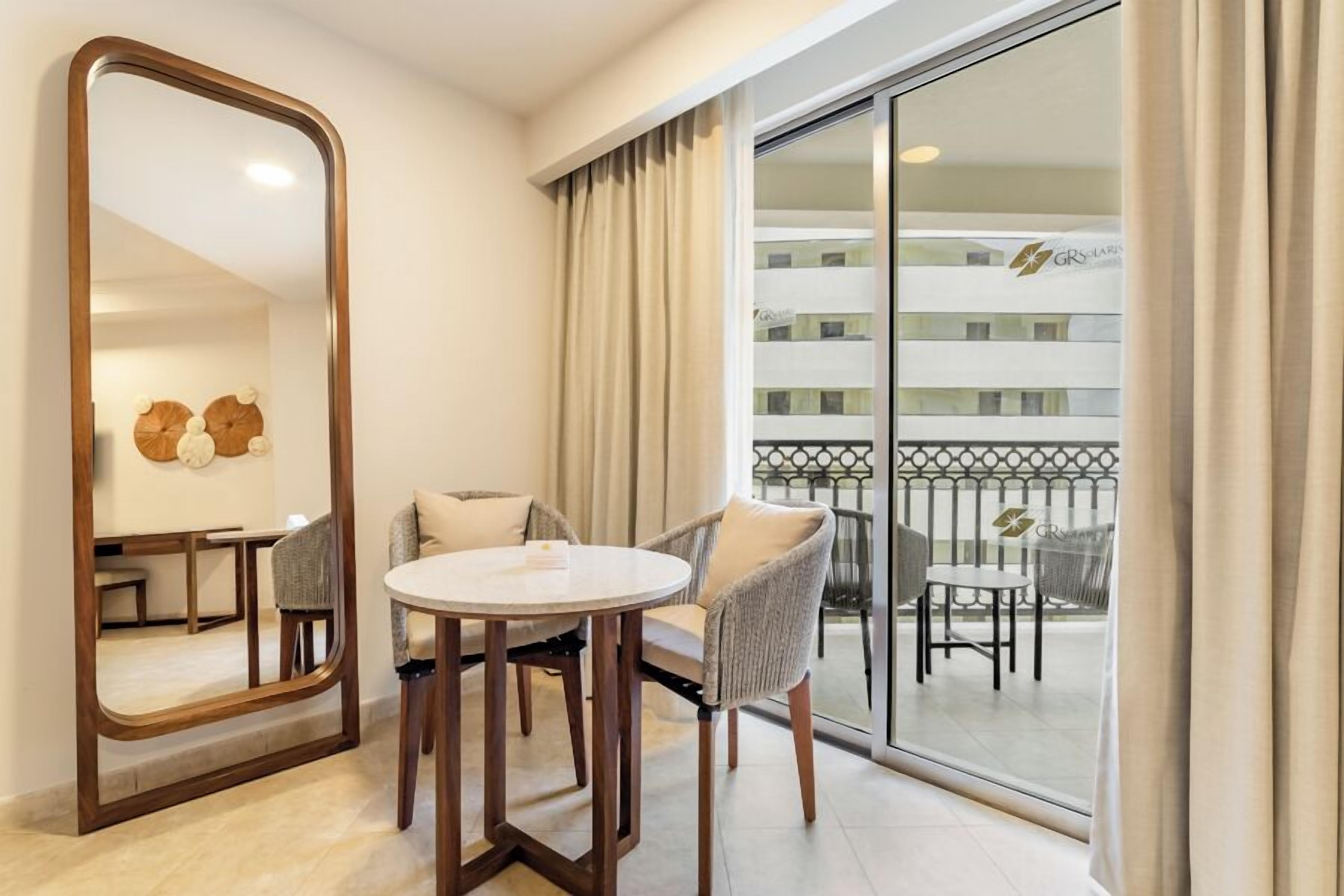 A hotel room with a large mirror, a round table, two chairs, a balcony with a railing, and a view of the hotel building.