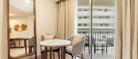 A hotel room with a large mirror, a round table, two chairs, a balcony with a railing, and a view of the hotel building.