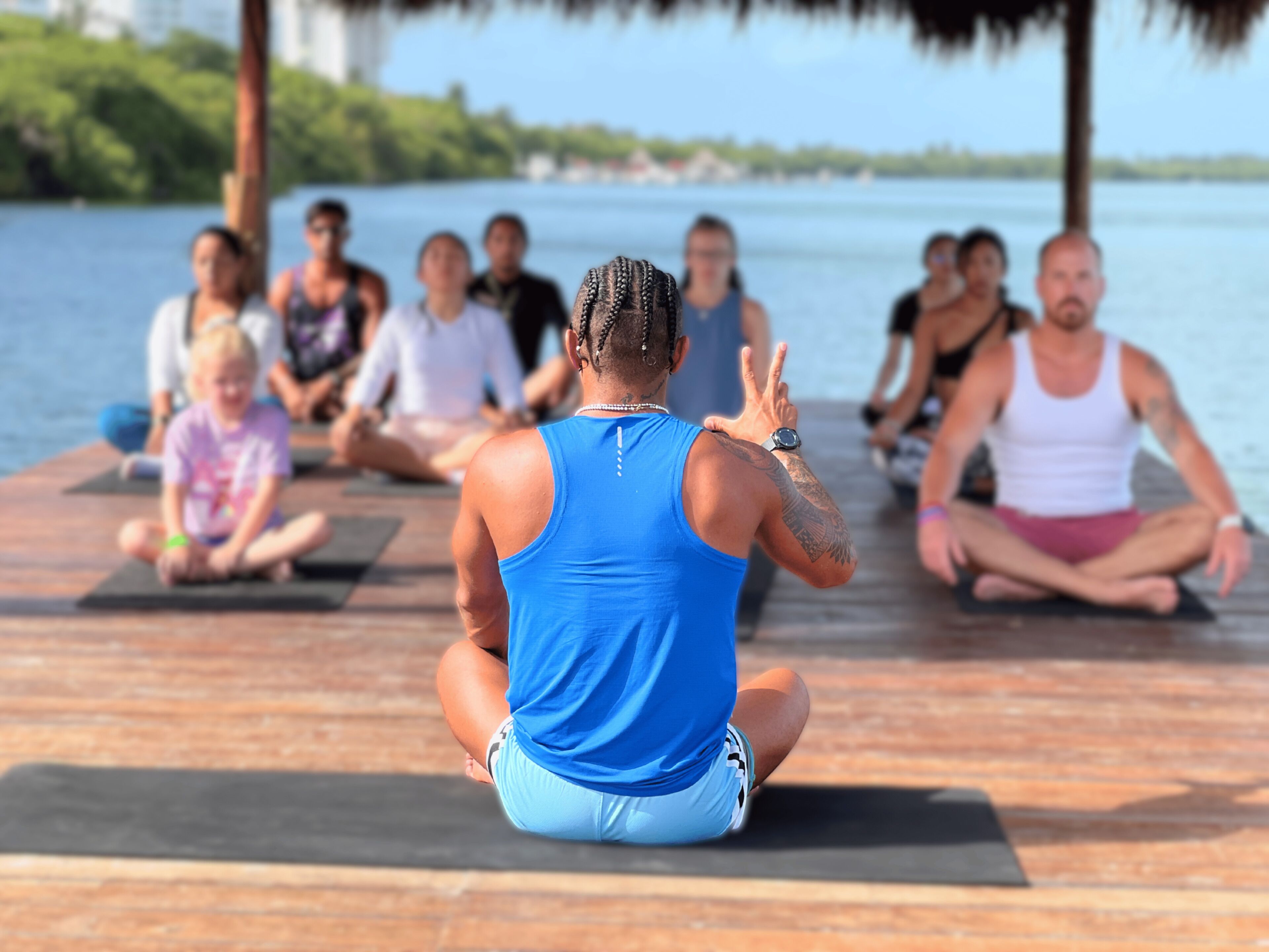 A group of people sitting on yoga mats in a semi-circle on a wooden deck by a body of water.