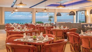 A restaurant with tables set for dining, large windows with a view of the sea, and hanging pendant lights.