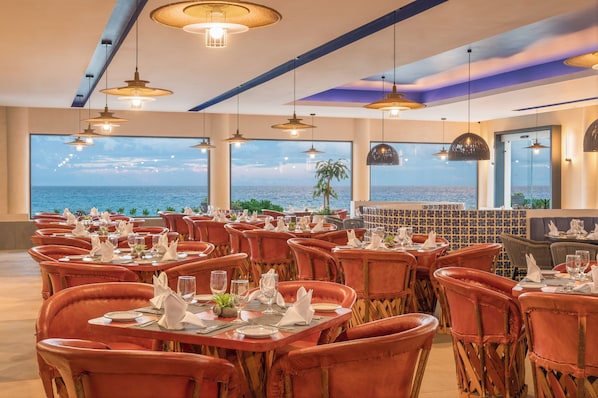 Un restaurant avec des tables dressées pour le service, de grandes fenêtres offrant une vue sur la mer et des lampes suspendues décoratives.