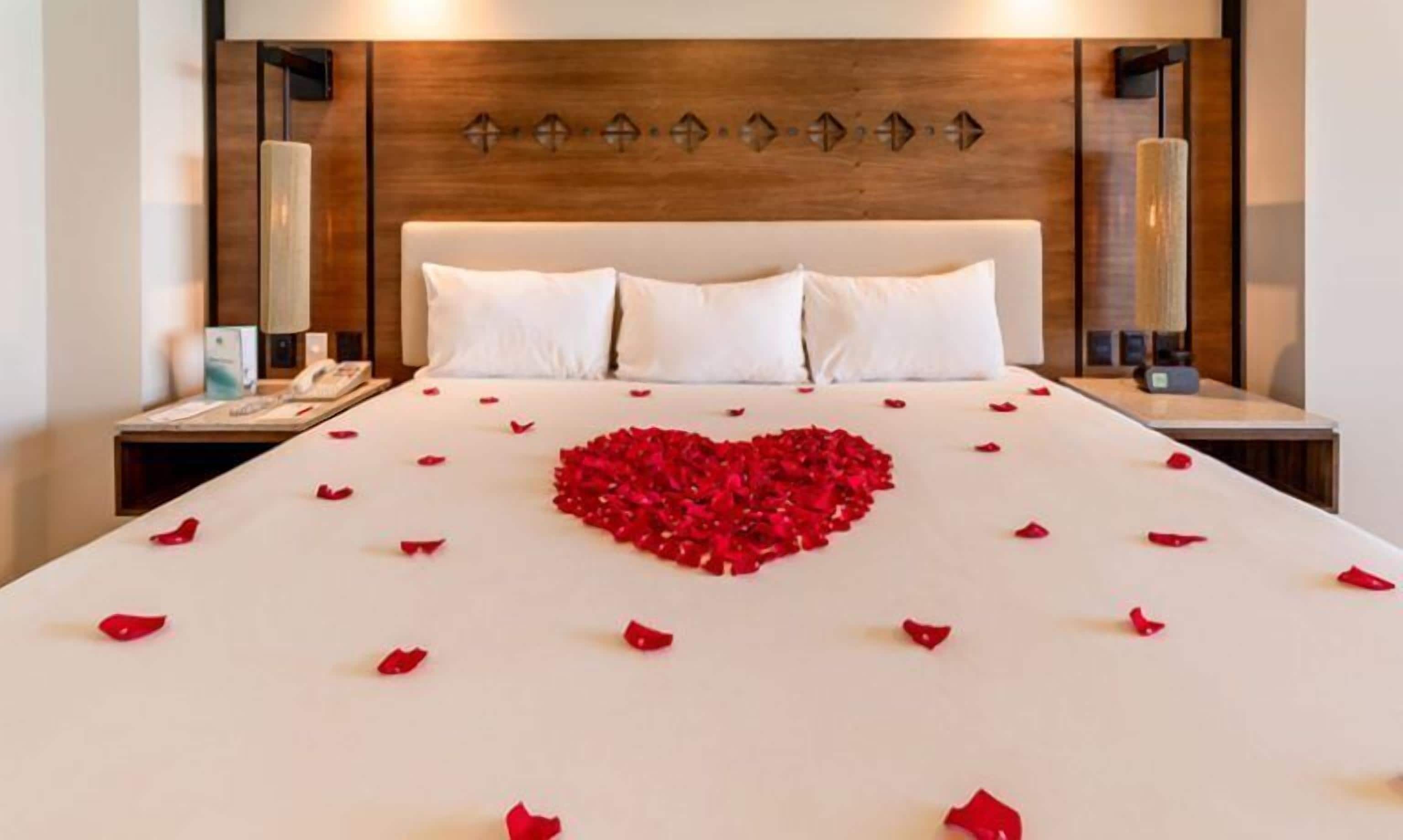 Une chambre d’hôtel avec un grand lit, une tête de lit en bois, des lampes de chevet et une décoration en forme de cœur faite de pétales de roses sur le lit.