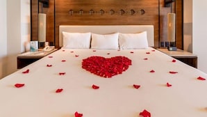 A hotel room with a large bed, a wooden headboard, bedside lamps, and a heart-shaped arrangement of rose petals on the bed.