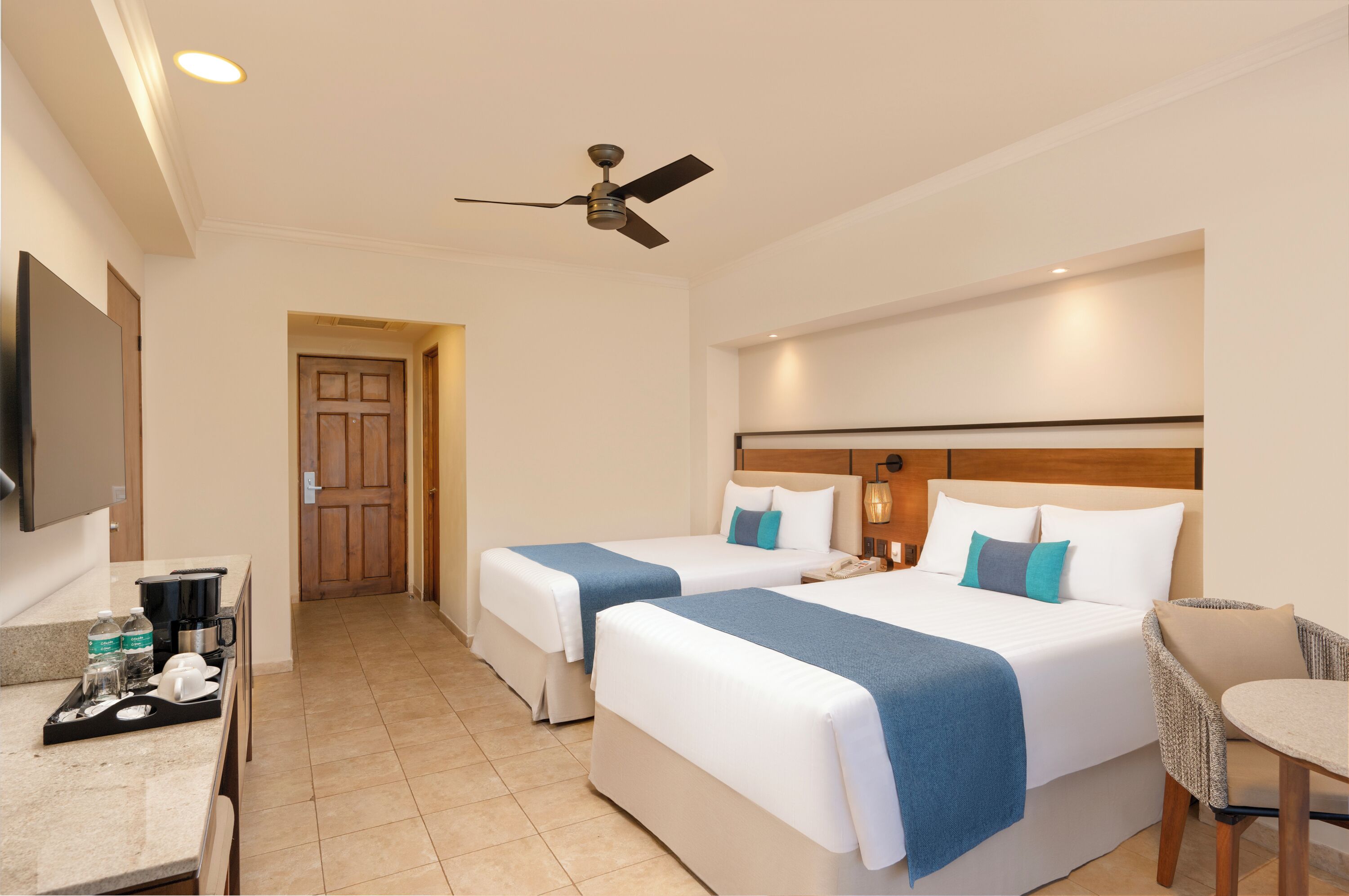 Quarto de hotel com duas camas, uma escrivaninha com cafeteira, televisão e ventoinha no teto.
