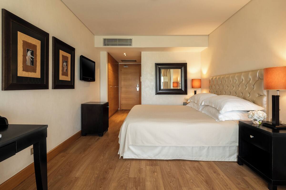 A hotel room with a bed, bedside tables, a desk, a flat-screen TV, framed artwork, and a mirror.