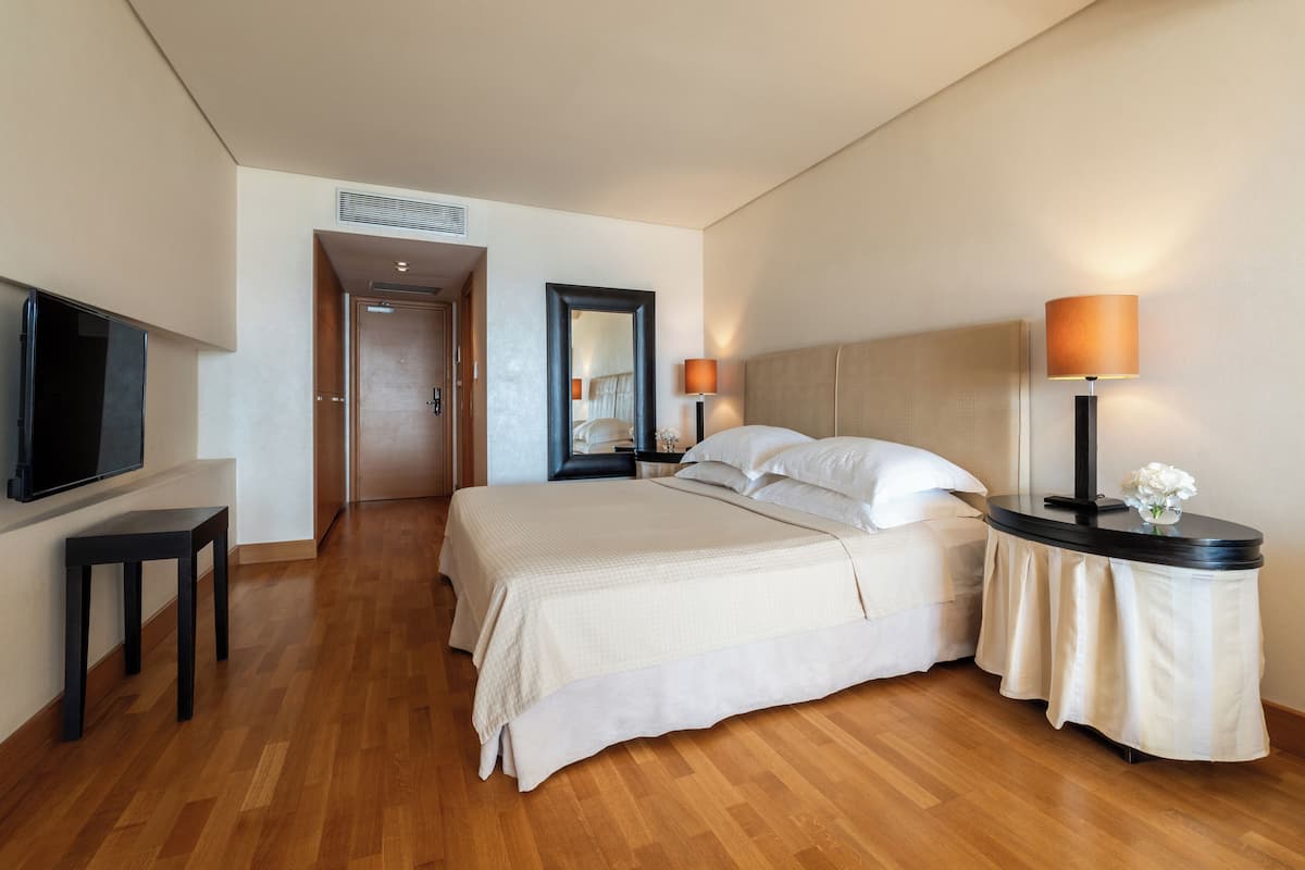 A hotel room with a large bed, bedside lamps, a TV, and a wooden floor.