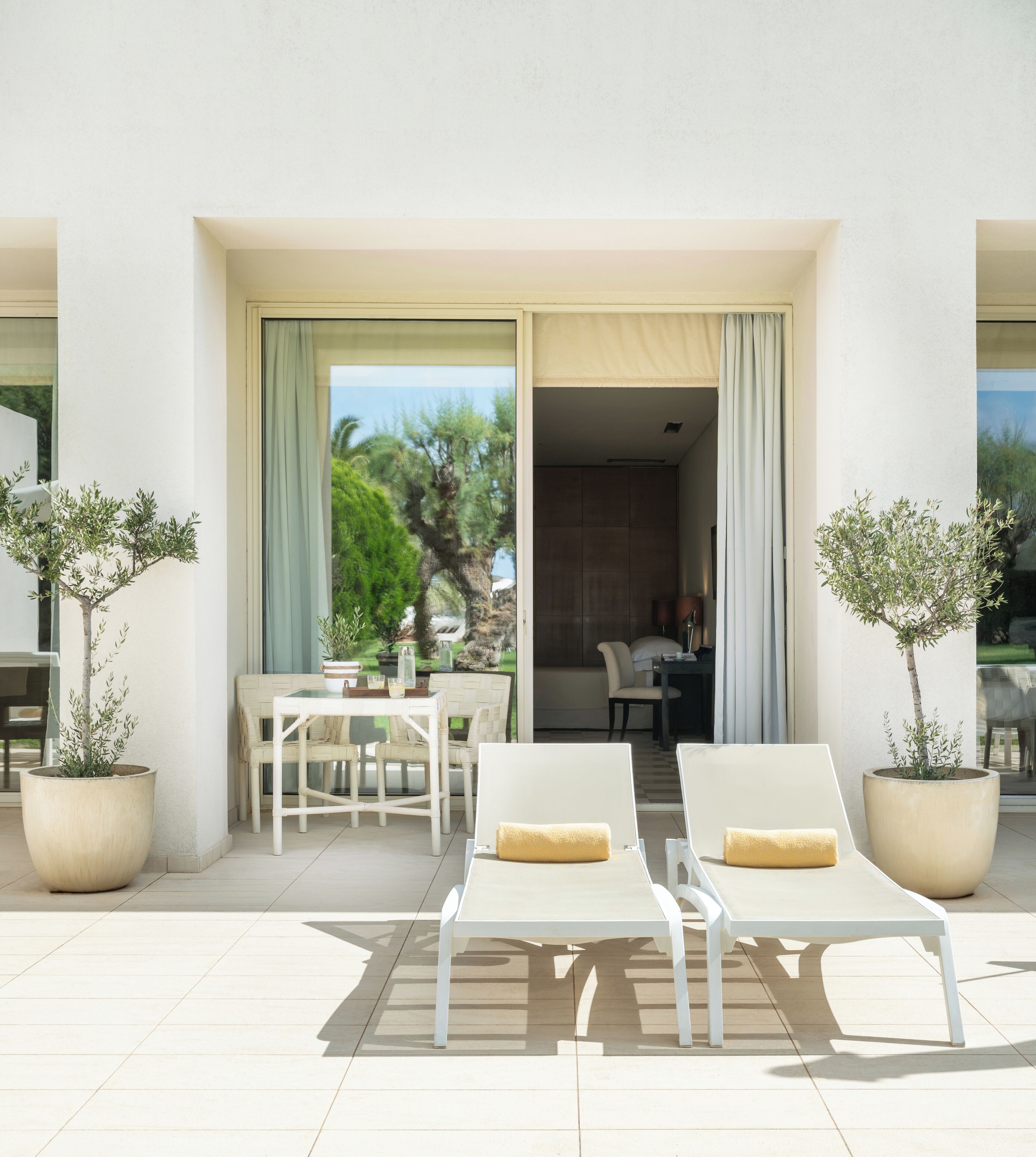 A modern outdoor seating area with white lounge chairs and a table, surrounded by large potted plants.