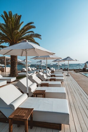 Un área de descanso frente a la playa con sillones blancos, mesas de madera, sombrillas que dan sombra y una palmera al fondo.
