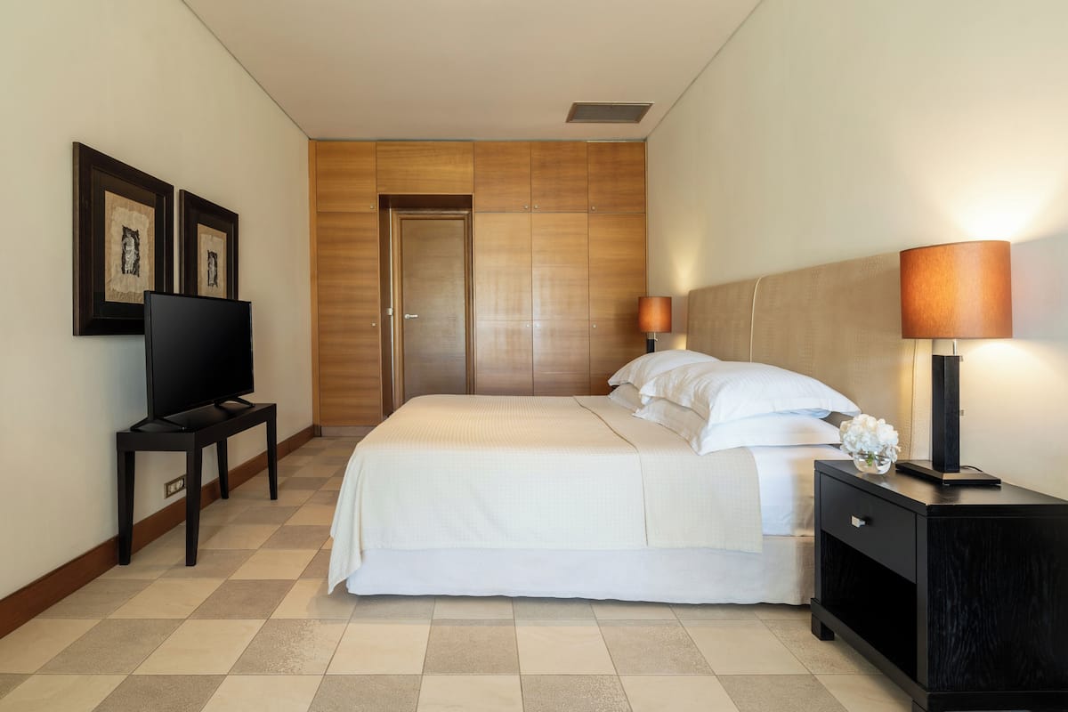 A hotel room with a bed, a TV on a stand, a nightstand with a lamp, and a door.