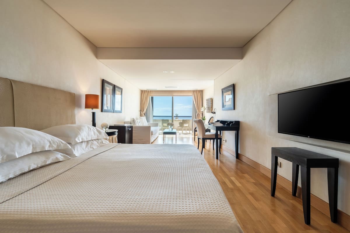 A hotel room with a large bed, a desk, a chair, a television, and a view of the sea.