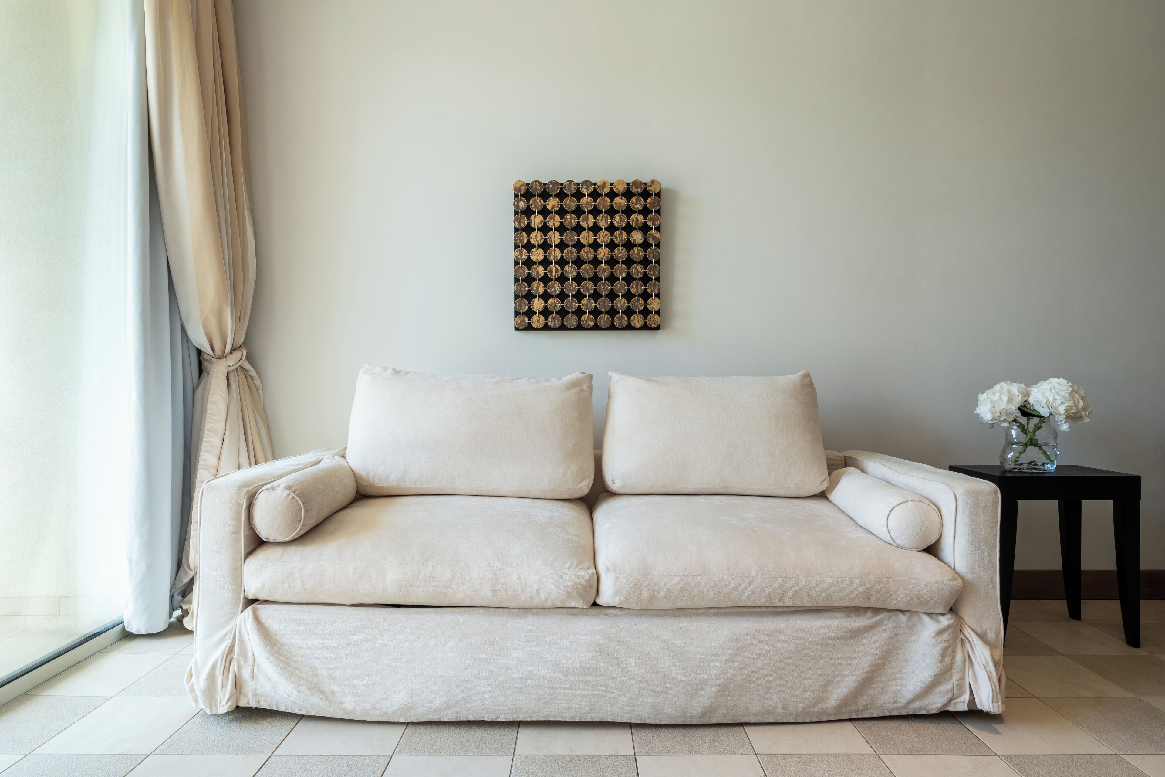 Ein beiges Sofa in einem Raum mit Fliesenboden, einem Wandbild und einem kleinen Tisch mit Blumen in einer Vase.