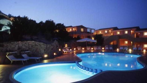 A swimming pool with lounge chairs and umbrellas, surrounded by a stone wall and a building with multiple windows. - Stelle Marine (Arzachena)