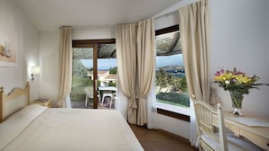 A bedroom with a large bed, a nightstand, a lamp, a window with curtains, a view of the outdoors, and a vase with flowers. - Stelle Marine (Arzachena)