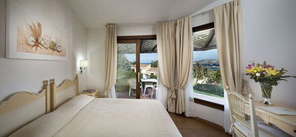 A bedroom with a large bed, a nightstand, a lamp, a window with curtains, a view of the outdoors, and a vase with flowers. - Stelle Marine (Arzachena)