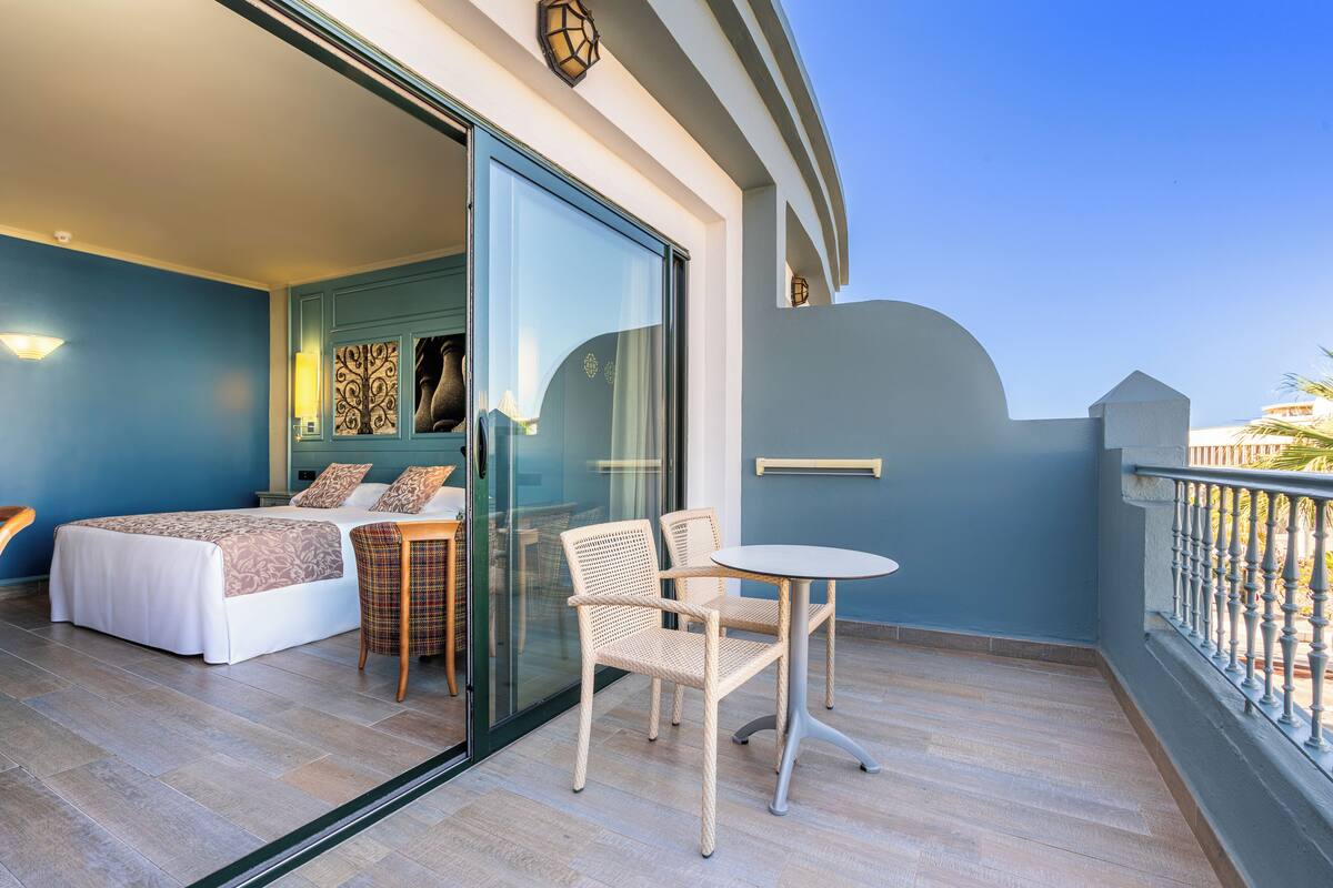 A balcony with a bed, two wicker chairs, a small round table, and a glass door leading to a room with a bed and a decorative wall.