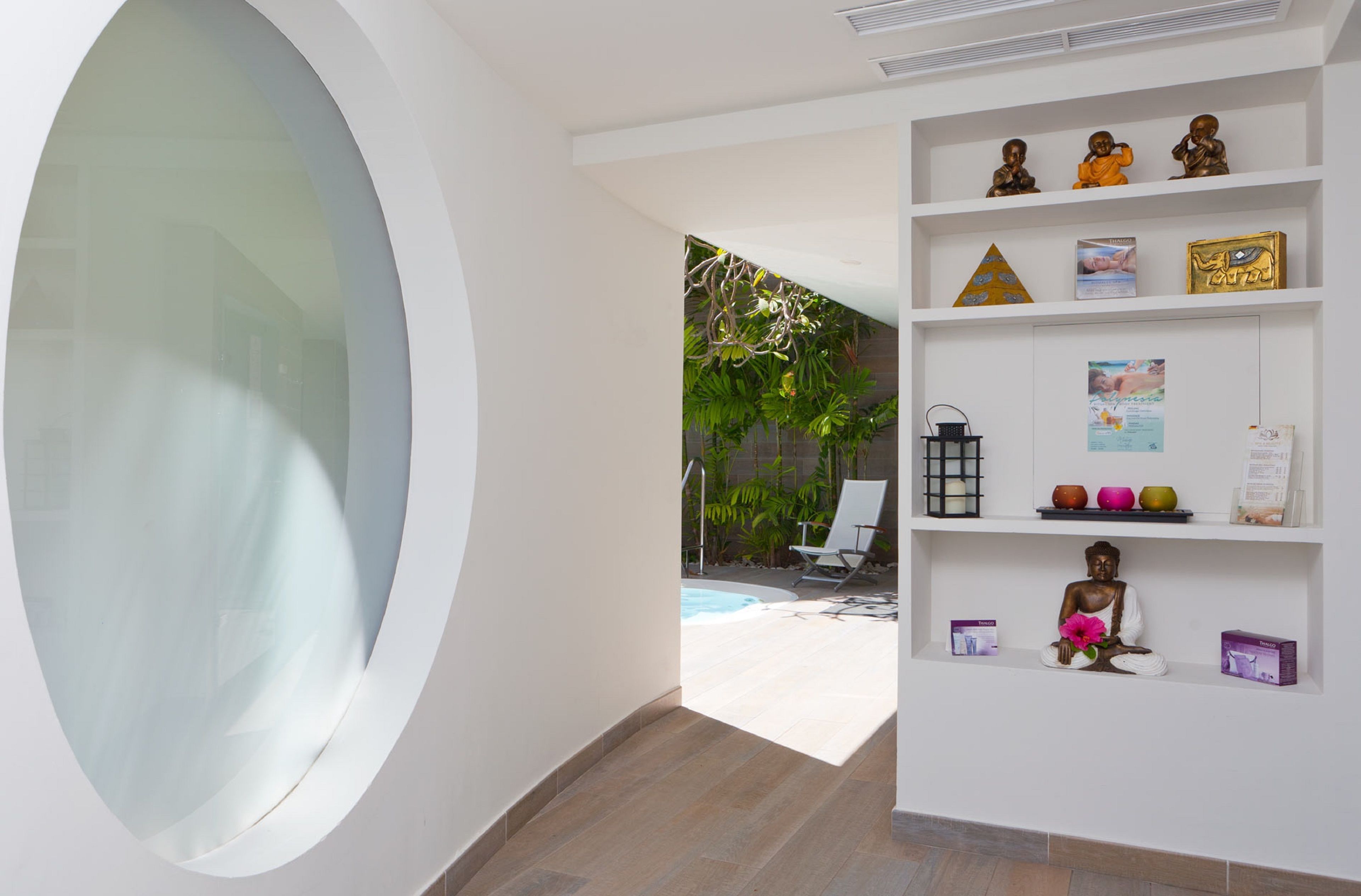 A modern interior with a large circular window, a white wall with shelves displaying various items, and a view of an outdoor area with a pool and plants.