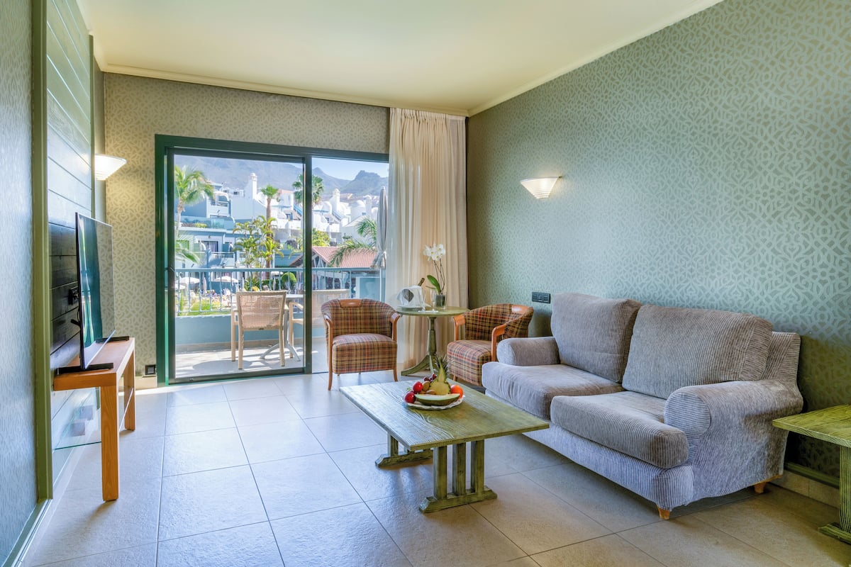 A living room with a sofa, two chairs, a coffee table with fruit, a TV, and a balcony with a view of buildings and palm trees.