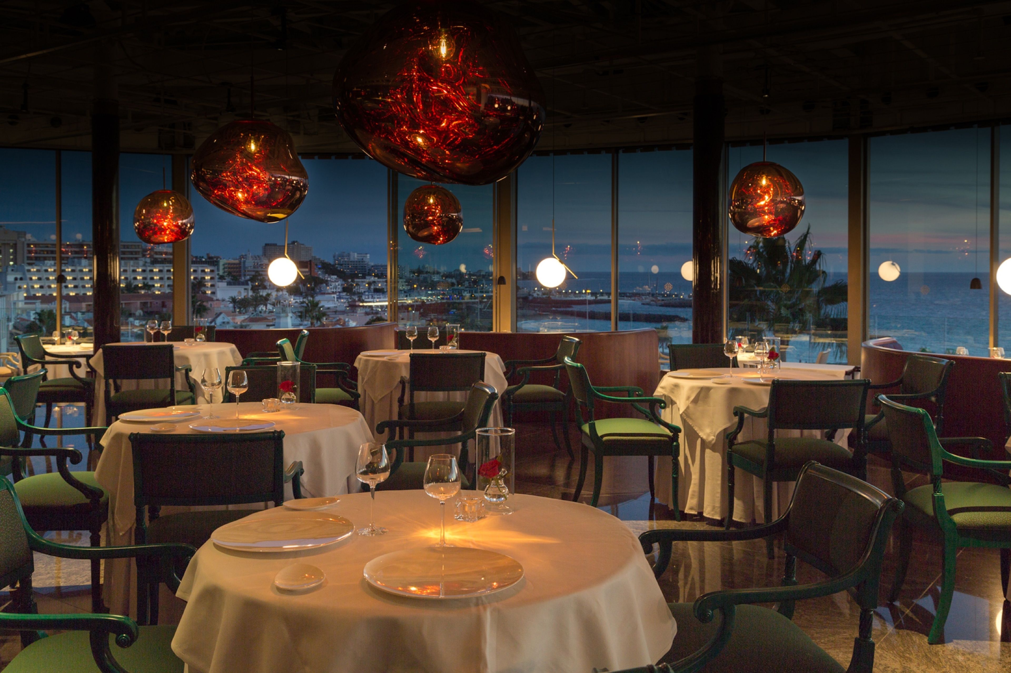 A restaurant with round tables, green chairs, and large windows offering a city view.