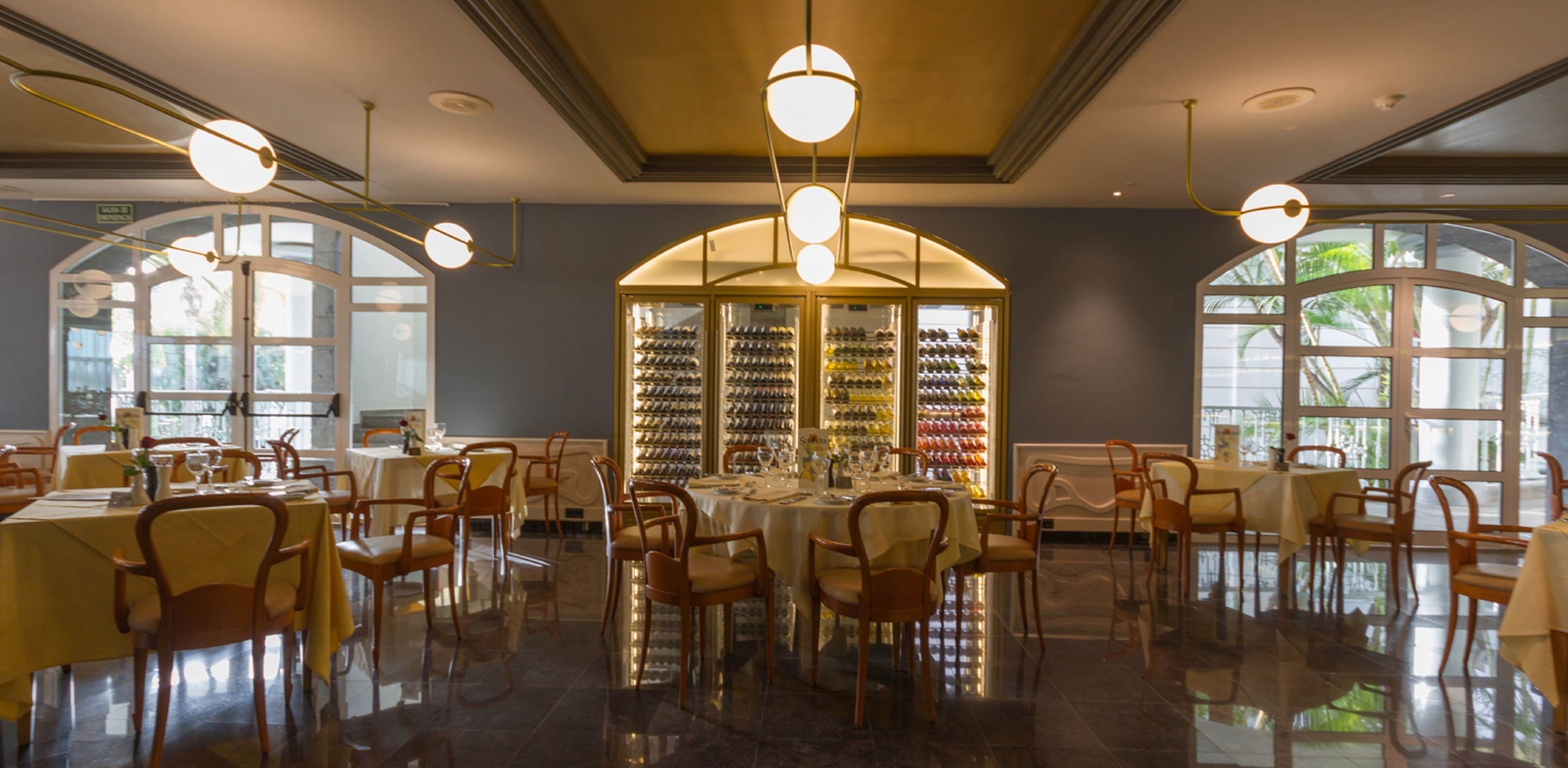 A restaurant with round tables and chairs, a wine rack in the background, and large windows.