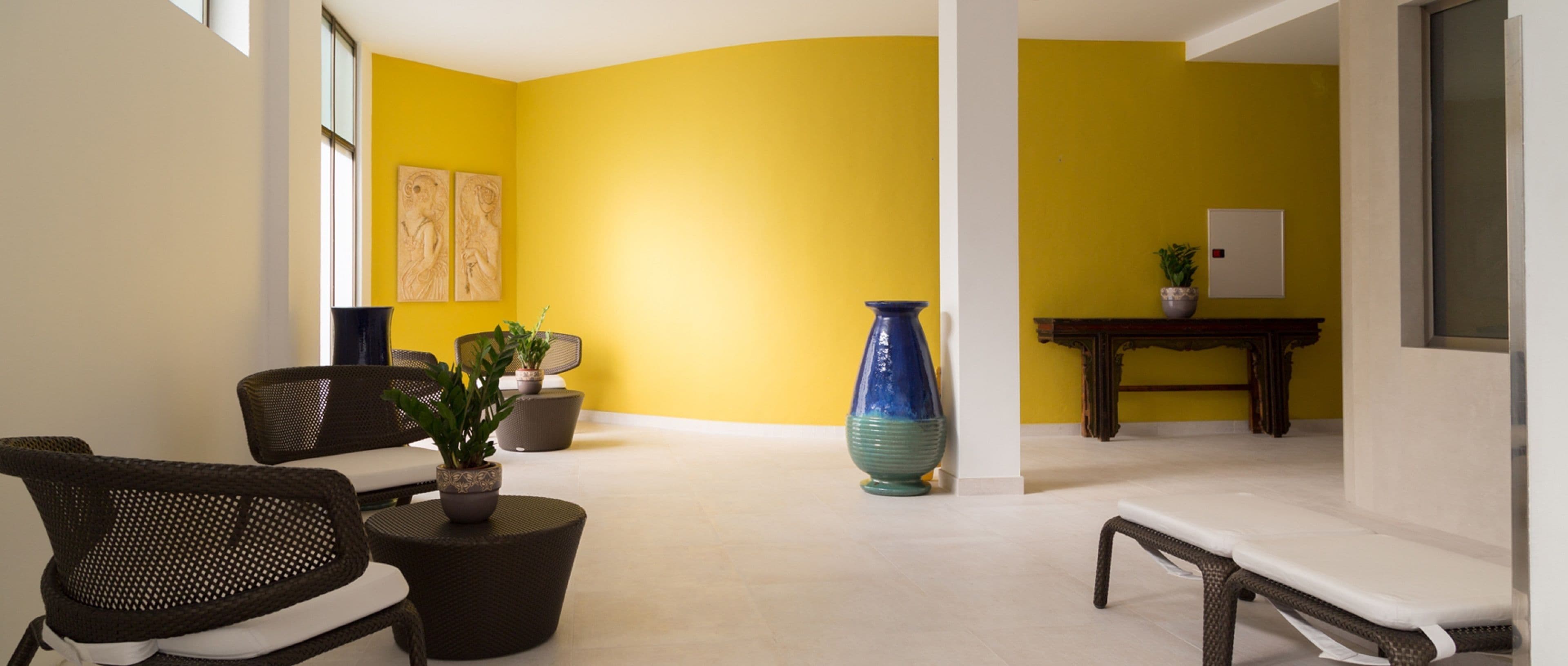 Una habitación con paredes amarillas, un jarrón azul grande, muebles de mimbre y un banco blanco.
