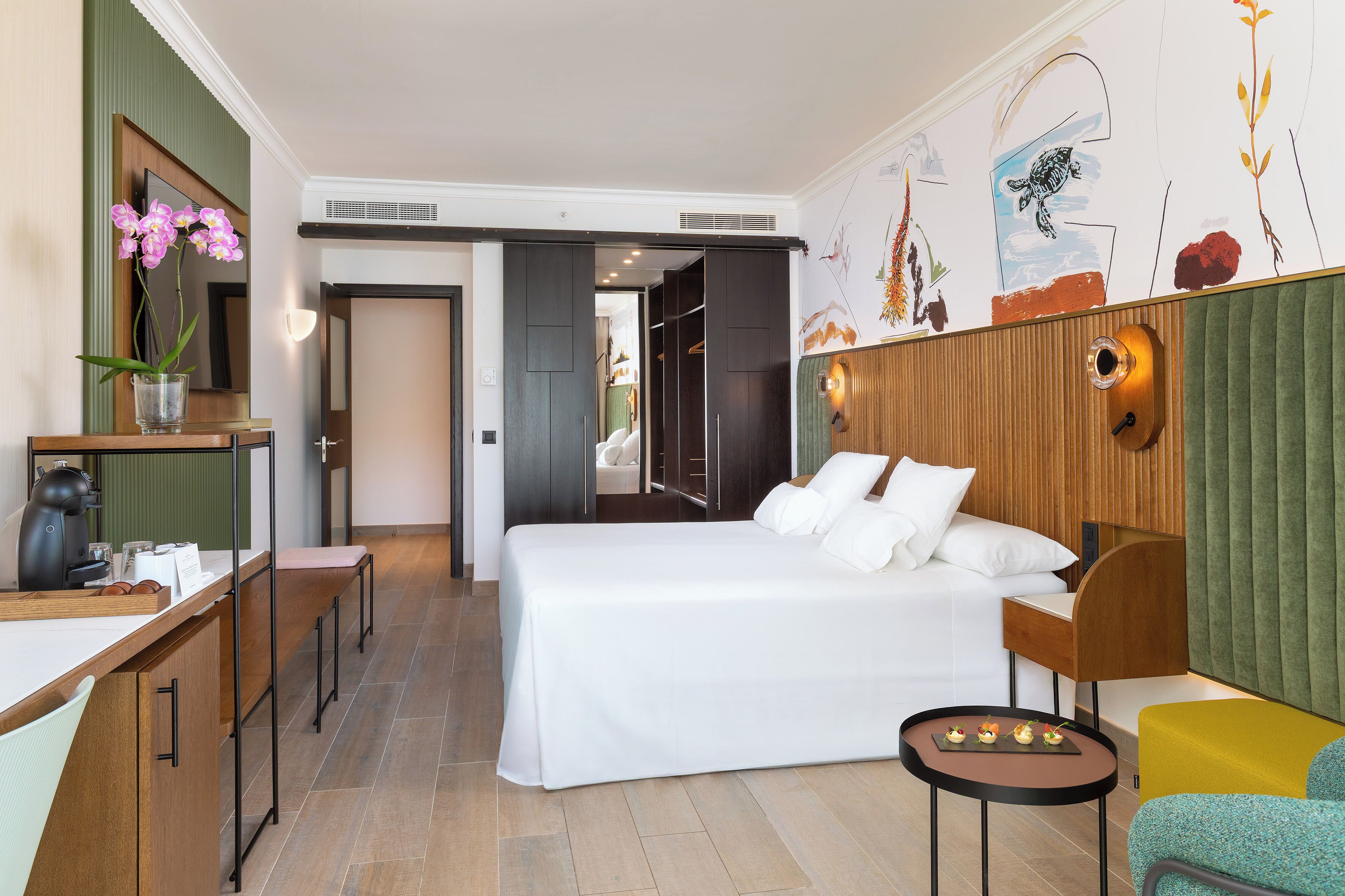 A modern hotel room with a large bed, a desk with a coffee maker, a small table with snacks, and a green upholstered armchair.