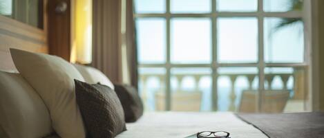 A neatly made bed with pillows, a book, and glasses on it, with a view of a balcony and a window in the background.