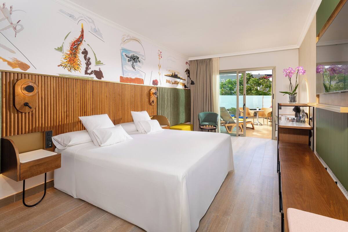 Ein modernes Hotelzimmer mit einem großen Bett, einem Holzkopfteil, einem kleinen Schreibtisch, einem Stuhl und Blick ins Freie.