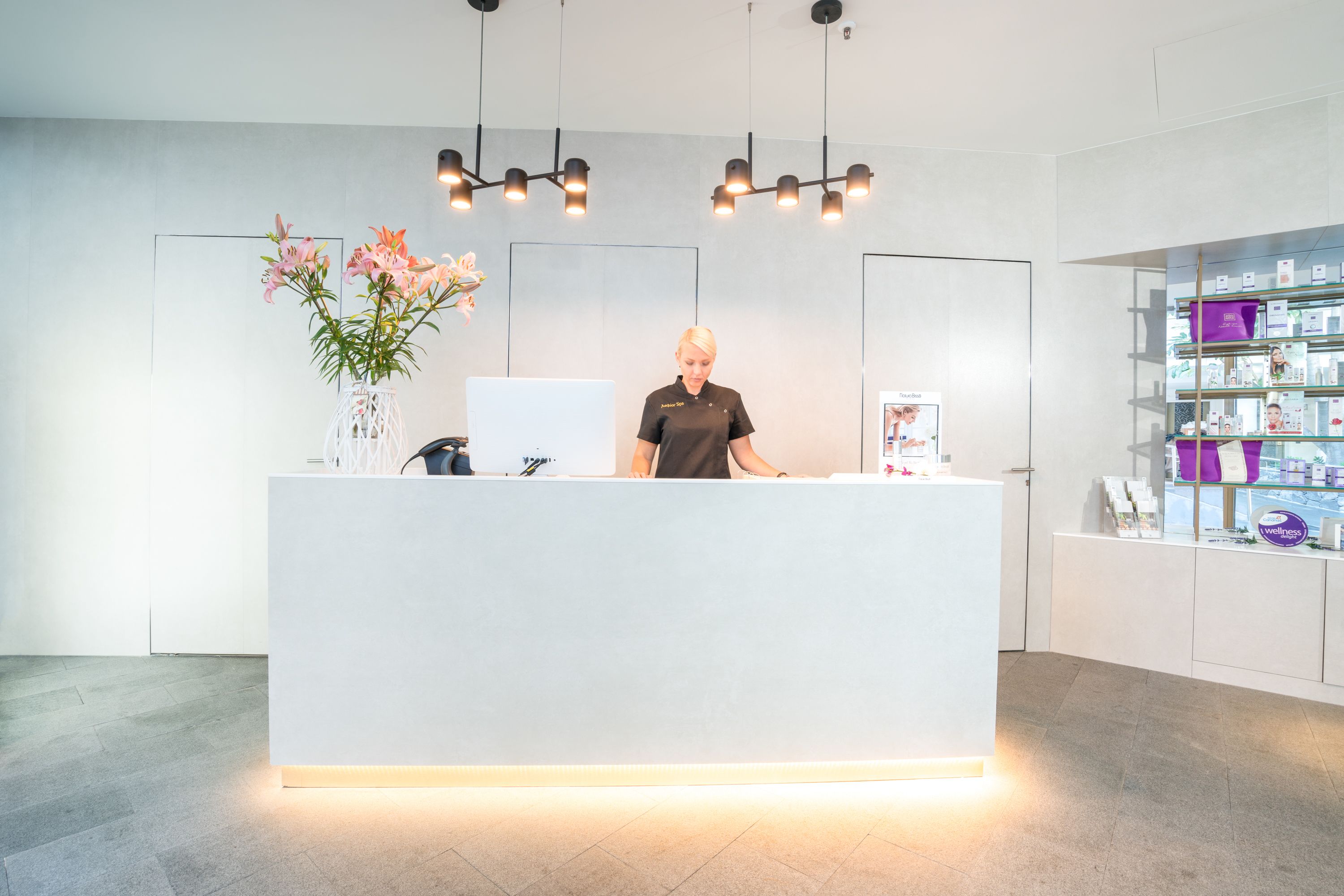 A reception desk with a white counter, a vase of pink flowers, and a person standing behind it.