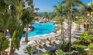 Área de piscina en un resort con sillones de descanso y sombrillas, rodeada de palmeras y vistas al océano.