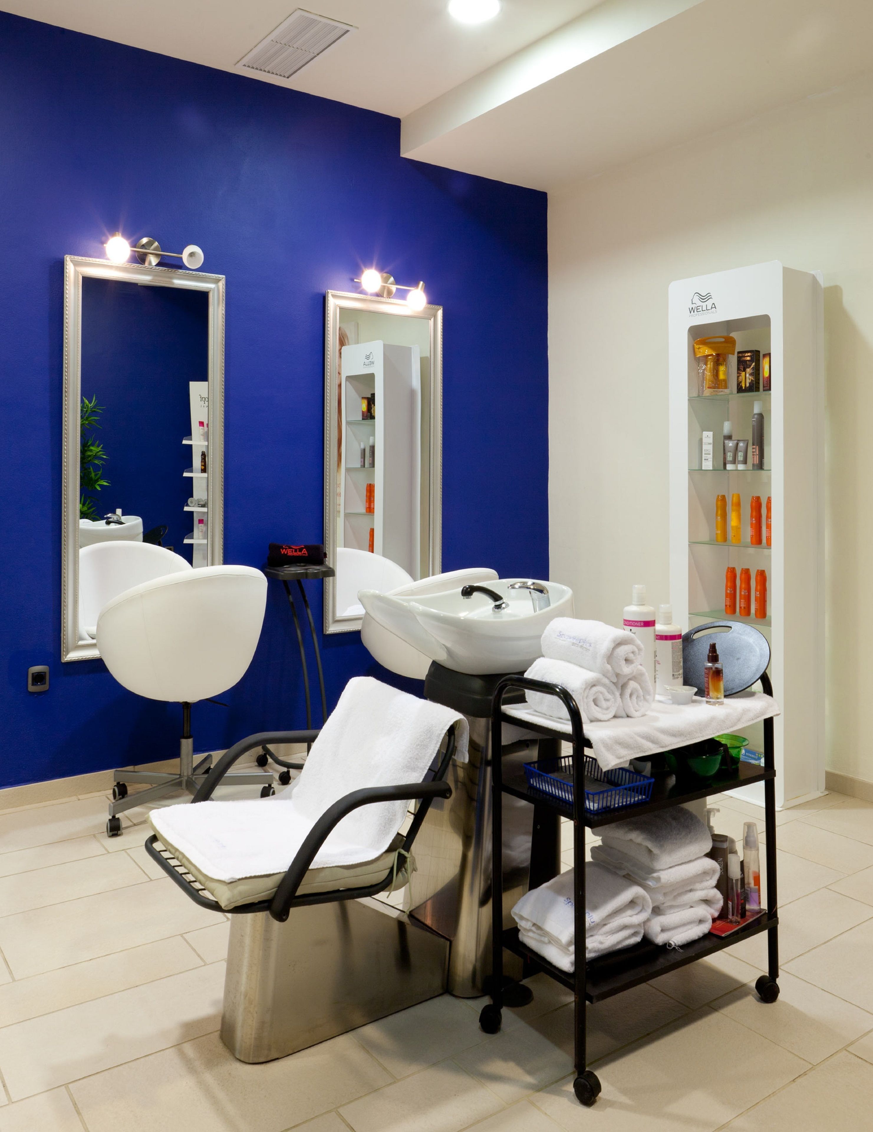 A modern bathroom with a blue accent wall, two large mirrors, a white sink, a chair, and a shelf with toiletries.