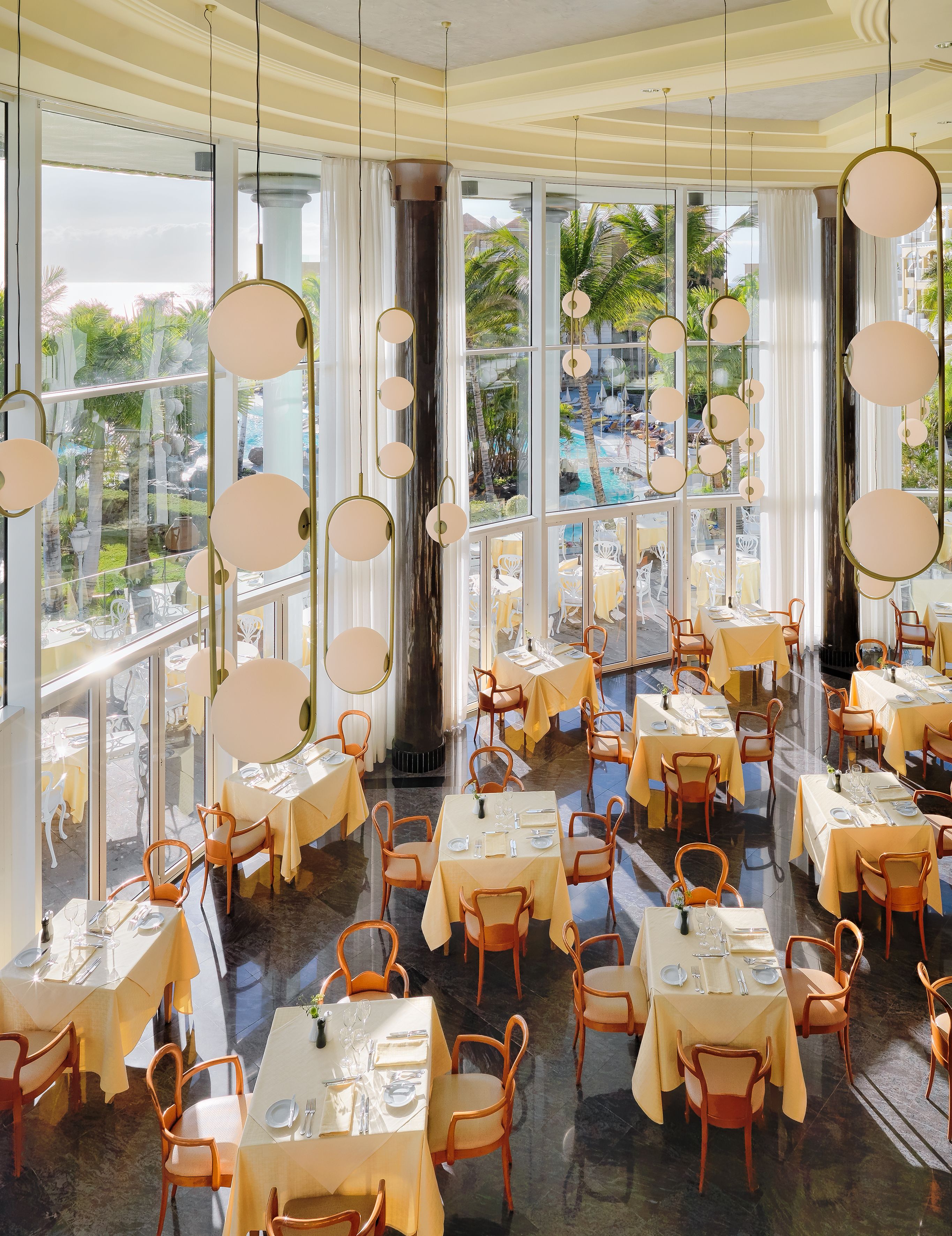 Un restaurante con varias mesas y sillas, ventanas amplias y lámparas colgantes.