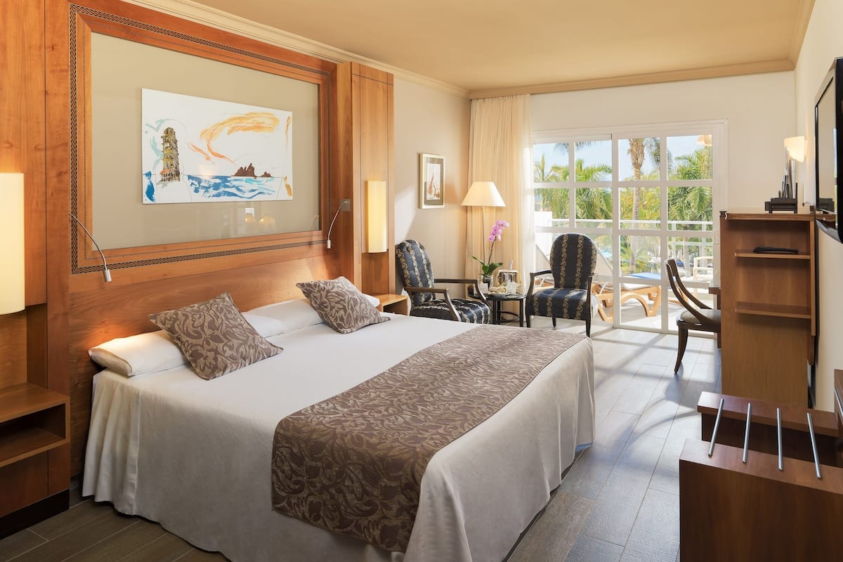 Ein Hotelzimmer mit einem großen Bett, einem Schreibtisch, einem Stuhl und einem Fenster mit Blick auf Palmen.