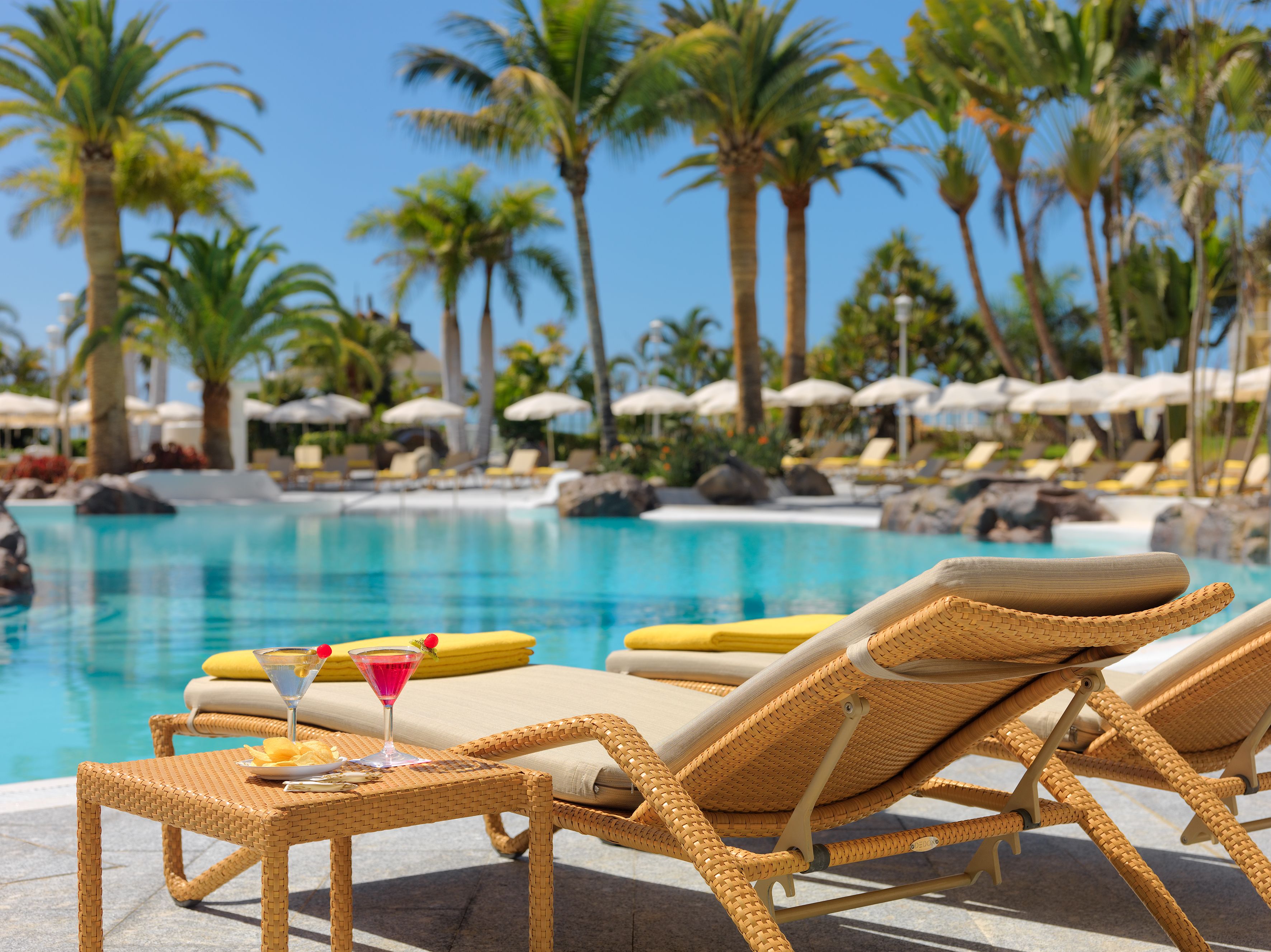 Un área junto a la piscina con sillones de mimbre, una mesa con una bebida y sombrillas.