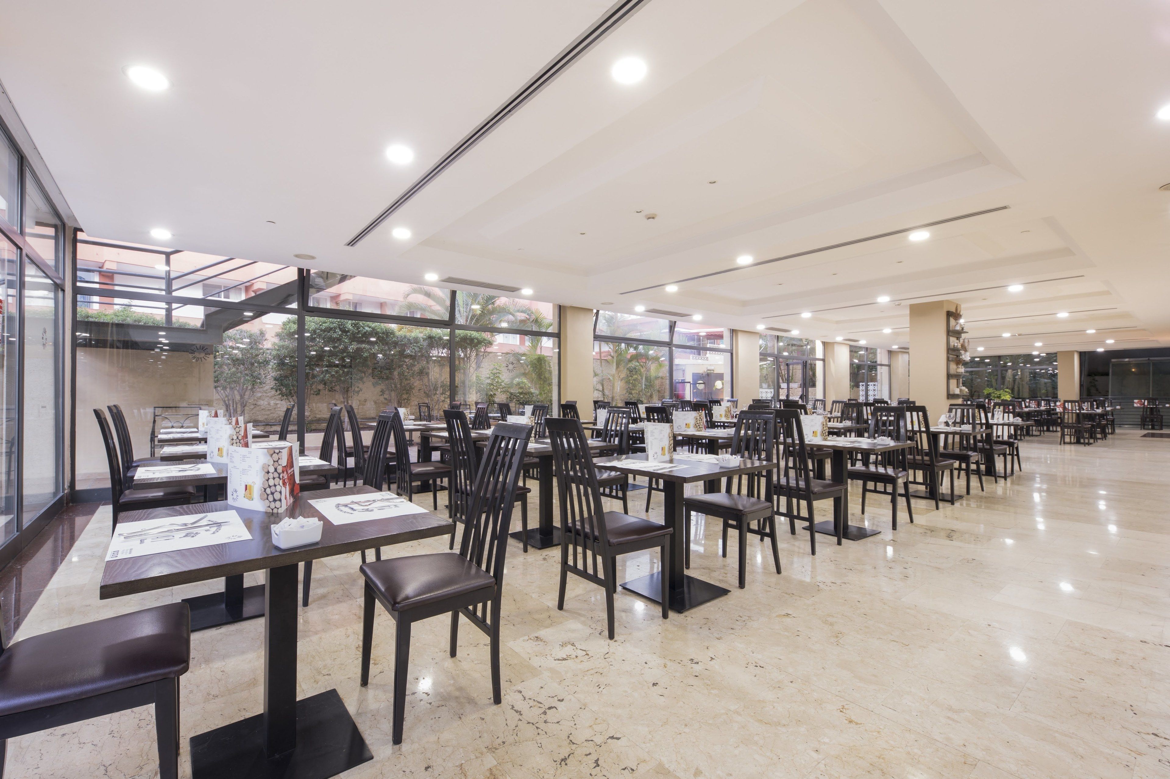 A spacious indoor dining area with multiple tables and chairs, large windows, and a high ceiling with recessed lighting.