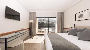 Habitación de hotel con cama, televisor montado en la pared, escritorio con silla y vista a la ciudad a través de un ventanal grande.