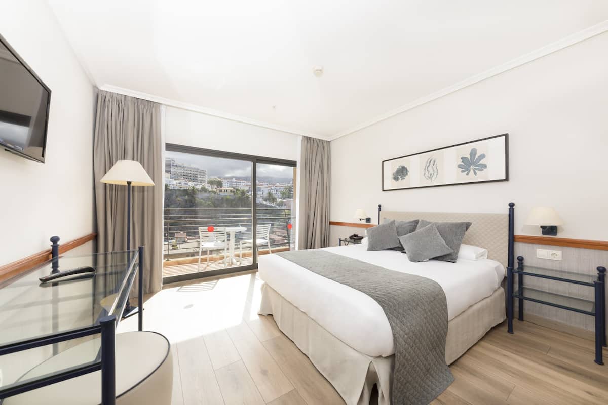 Une chambre d’hôtel avec un grand lit, un balcon avec une table et des chaises, et une vue sur la ville.