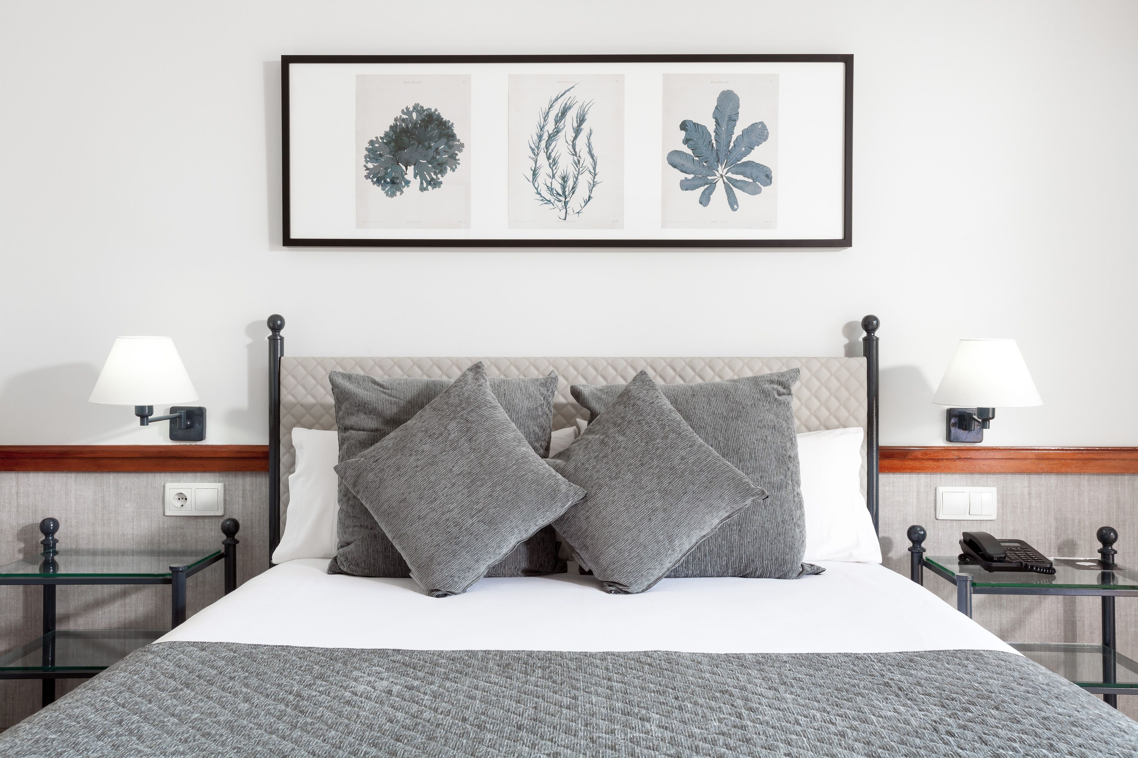 A neatly made bed with gray bedding and pillows, a framed botanical print above it, and bedside tables with lamps and a phone.