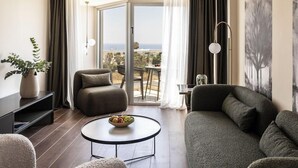 A modern living room with a sofa, armchair, and a round coffee table with fruit. There is a balcony with a view of the city and a painting on the wall. - Crown Resorts Horizon Hotel (Pegeia)