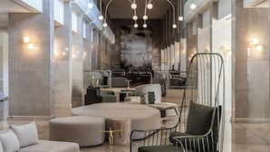 A spacious lobby with a modern design, featuring a central seating area with ottomans and chairs, a large abstract mural on the wall, and a staircase leading to upper floors. - Crown Resorts Horizon Hotel (Pegeia)