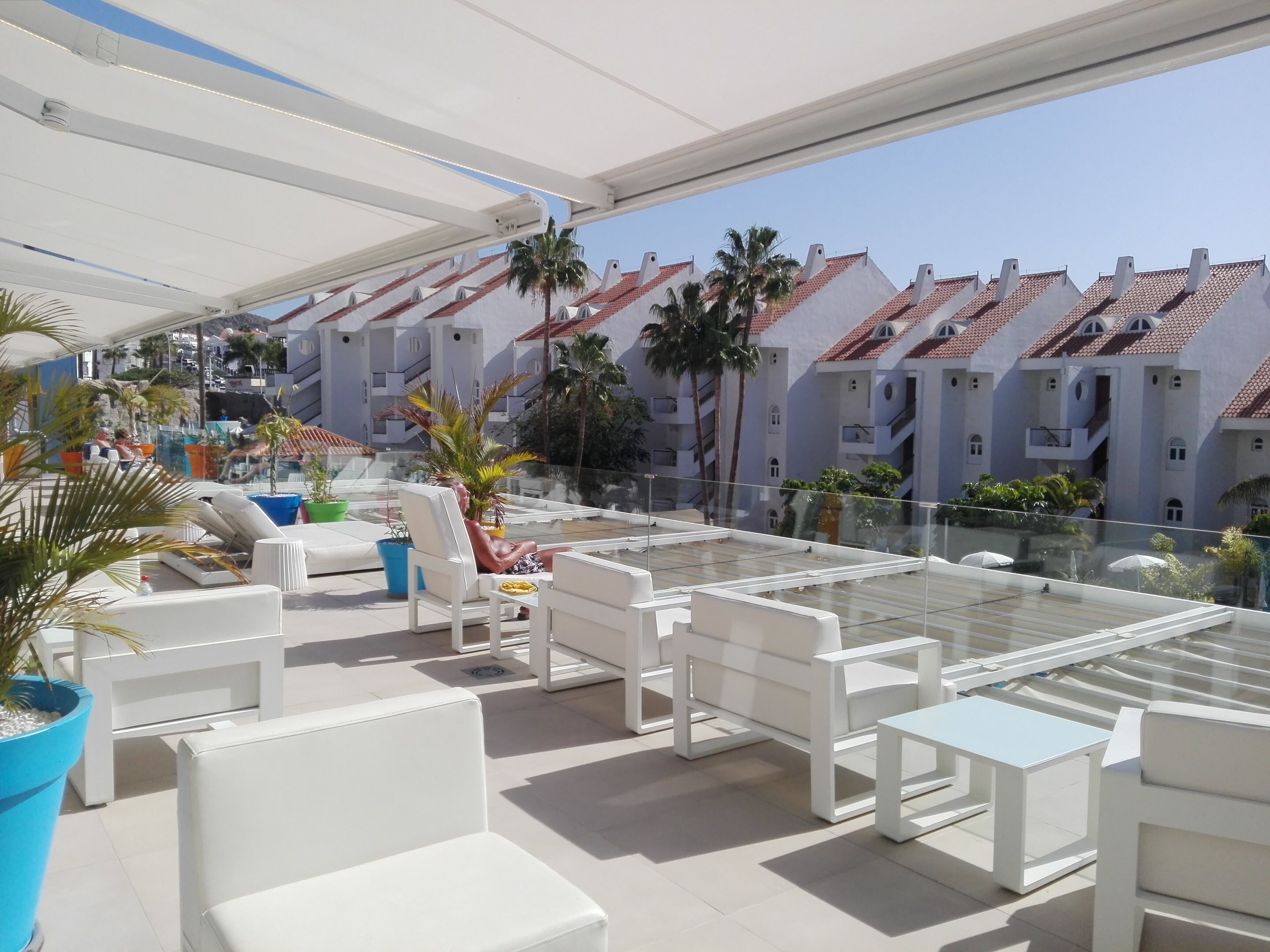 A rooftop terrace with white furniture, palm trees, and a view of buildings with red-tiled roofs.