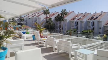 A rooftop terrace with white furniture, palm trees, and a view of buildings with red-tiled roofs.