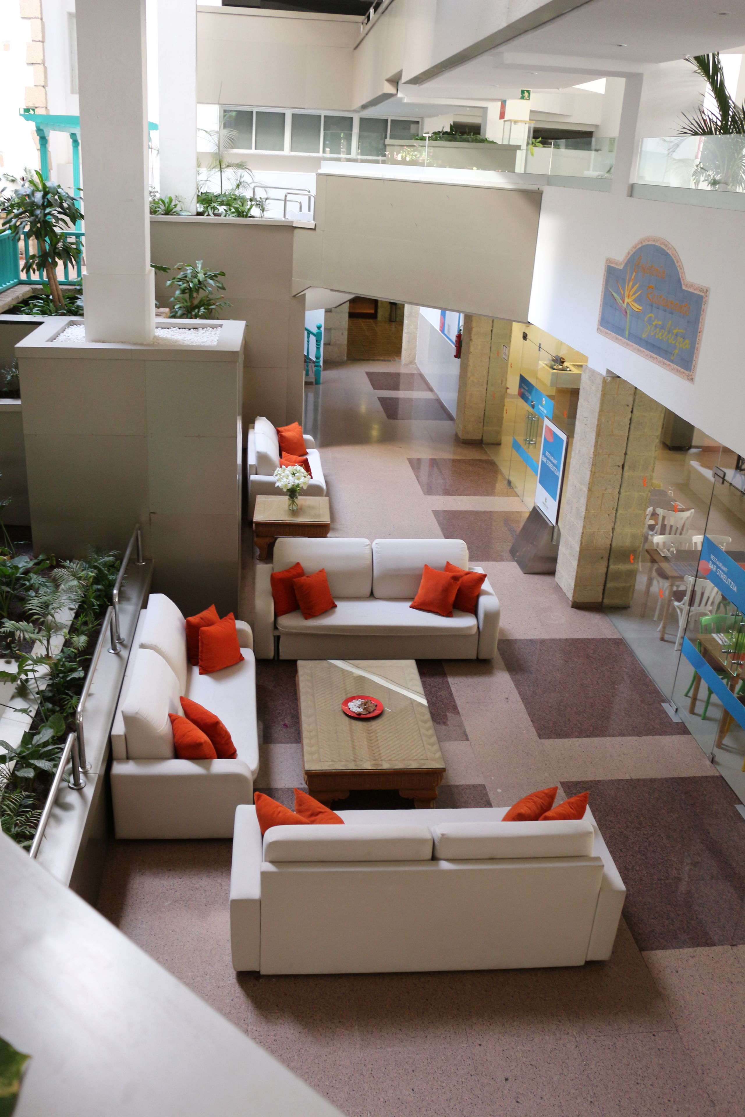 A spacious indoor area with white sofas and orange cushions, a coffee table, and potted plants.