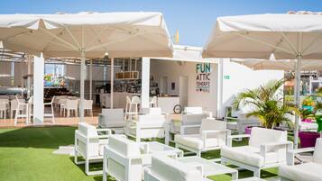 Mobiliario exterior con sillas blancas y sombrillas, zona de bar y un cartel que dice "FUN ATTIC TENERIFE".