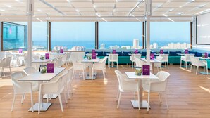 A spacious indoor area with tables and chairs, large windows offering a city view, and a high ceiling with a grid pattern. - Paradise Park Fun Lifestyle Hotel (Arona)