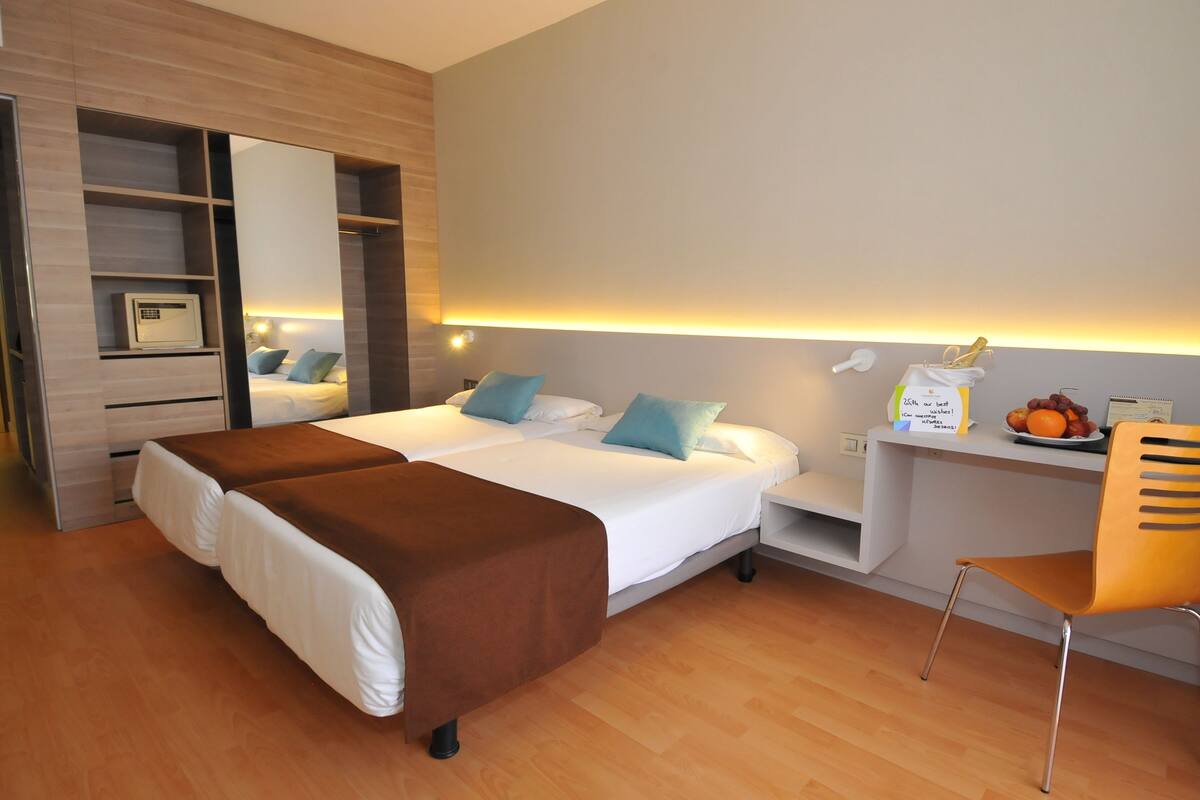 Habitación de hotel con cama doble, escritorio con silla, armario y mesita de noche con frutero.