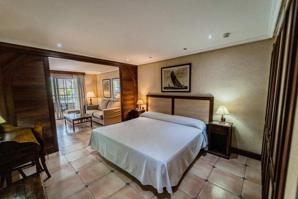 Una habitación de hotel con una cama grande, un escritorio, una silla, una mesita de noche con lámpara y un cuadro en la pared.