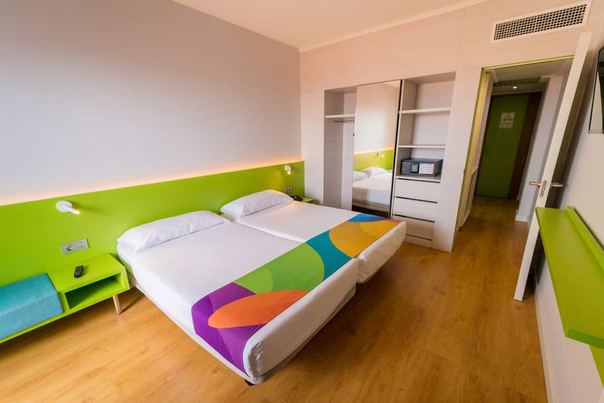 Una habitación de hotel moderna con una cama, una mesita de noche verde, un espejo y un armario.